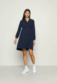 Tengerész stílusú, hosszú ujjú, V-kivágású, aszimmetrikus hemű ruha, puha anyagból készült. Fehér sportcipővel viselve.