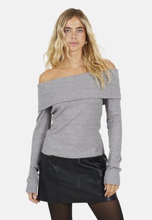 Jonge vrouw die een grijze off-shoulder geribbelde trui en een zwarte leren minirok draagt, staand tegen een witte achtergrond.