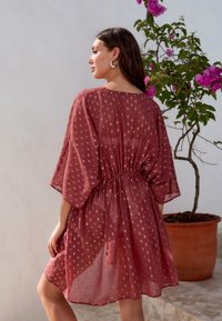 Priehľadné šaty hrdzavej farby so zúžením v páse, zvonovými rukávmi a jemným vzorom. Majú hlboký výstrih do V a voľný, plynulý strih.