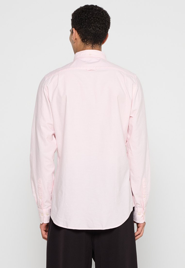 Eine Person mit lockigem Haar trägt ein langärmeliges, hellrosa Hemd, betrachtet von hinten, mit schwarzen Hosen und einem neutralen Hintergrund.