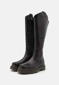 Black leather knee-high boots with a rounded toe, chunky tread sole, yellow stitching accents, and a zipper on the side.