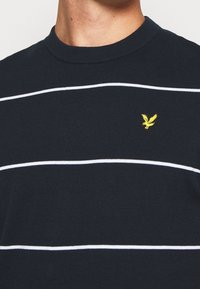 Pull à manches courtes bleu marine avec des rayures horizontales blanches et un petit logo aigle jaune sur la poitrine. Col rond côtelé. Matière en mélange de coton.