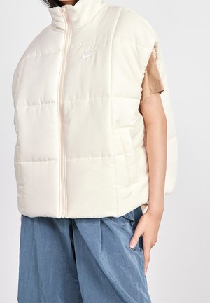 Persoon die een mouwloos wit gewatteerd Nike-vest draagt over een bruin kortermig shirt en losse blauwe broek, tegen een effen achtergrond.