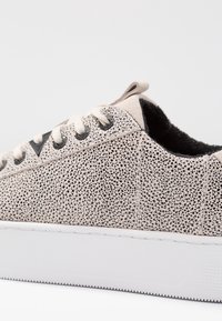 Baskets beiges avec une tige texturée et tachetée, une semelle blanche et des lacets ronds. Elles comportent une doublure en tissu et des accents noirs près des œillets.