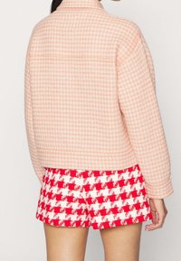 Růžový kabát s houndstooth vzorem a límečkem, s texturovaným povrchem, spojený s červenými a bílými kostkovanými šortkami. Zobrazen pohled zezadu.