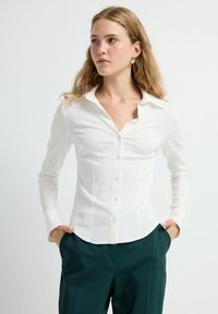 Witte slim fit blouse met een kraag, lange mouwen en een gestructureerde stof. Gekleed met groene broek, wat een slanke silhoutte laat zien.