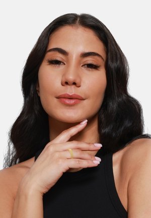 Anello in mesh d'oro indossato sull'indice, texture opaca, design delicato. La modella ha i capelli scuri e ondulati e un trucco dai toni neutri, con un top senza maniche.