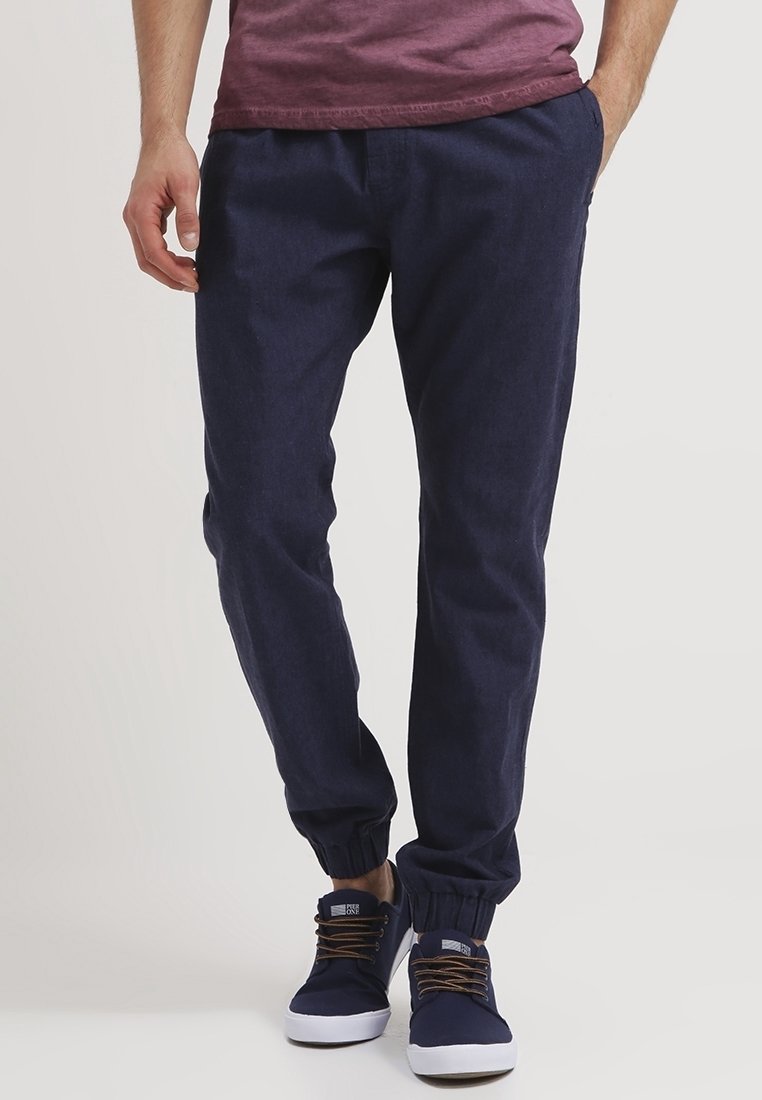 Man in marineblauwe joggers met elastische manchetten en marineblauwe sneakers met witte zolen en bruine veters.