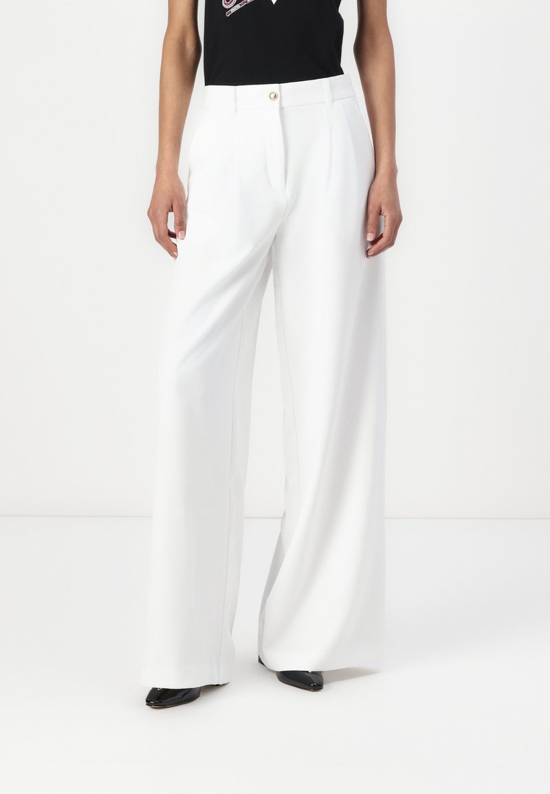 Femme portant un pantalon blanc à jambes larges et des chaussures noires pointues, associé à un top noir rentré, sur un fond blanc uni.