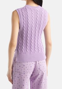 Liivinätön lilakuvioinen neulepaita yhdistettynä vaaleanpunaiseen kukkakuosiset farkkuihin, näkyy takaa henkilön päällä, jolla on pitkät tummat hiukset.