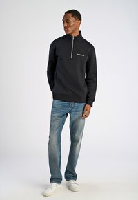 Felpa nera con zip e colletto alto. Presenta un logo bianco sul petto. Indossata con jeans azzurri e sneaker bianche.