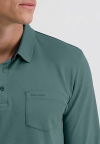 Dunkelgrünes Poloshirt aus glattem Stoff, mit Kragen, Knopfverschluss und einer linken Brusttasche, die mit "pierre cardin" geprägt ist.