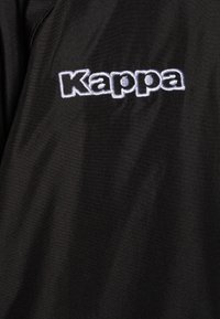 Veste de sport noire avec un logo "Kappa" brodé blanc bien en vue sur la poitrine. Tissu lisse et léger avec une légère brillance.