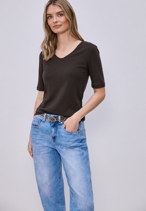 Mujer de pie con una mano en el bolsillo de sus jeans azul claro, vistiendo una blusa marrón oscuro de manga corta con escote en V y un cinturón con estampado de leopardo.