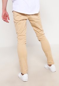 Persona che indossa pantaloni beige slim-fit, sneakers bianche e una camicia bianca, in piedi con le gambe leggermente divaricate su uno sfondo neutro.