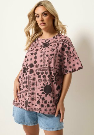 Ung kvinde med langt blondt hår iført en løs lyserød T-shirt med sorte sol- og blomsterprint og lyseblå denimshorts.