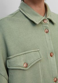 Lichtgroen button-down shirt met een kraag en een klepzak op de borst, getoond op de bovenkant van een persoon.