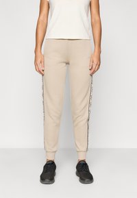 Μπεζ φόρμες με στενή εφαρμογή, που διαθέτουν μαύρες πλαϊνές ρίγες με σχέδιο λογότυπου. Φτιαγμένες από μαλακό βαμβακερό μίγμα.