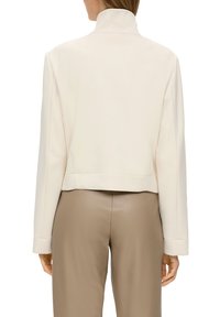 Geknipte, crèmekleurige jas met een hoge kraag, lange mouwen en een gladde textuur, gecombineerd met beige broeken. Schone lijnen en minimaal design.