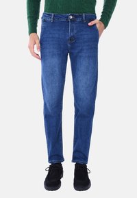 Jeans in denim di colore blu scuro con una silhouette dritta, dotati di cinque tasche e chiusura con bottone in metallo. Indossati con scarpe nere.