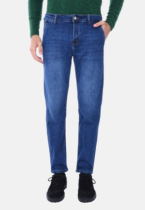 Jeans in denim di colore blu scuro con una silhouette dritta, dotati di cinque tasche e chiusura con bottone in metallo. Indossati con scarpe nere.