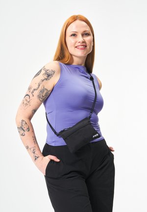 Roodharige vrouw met paarse mouwloze top, zwarte broek, klein zwart schoudertasje en meerdere tatoeages op haar linkerarm, glimlacht zelfverzekerd.