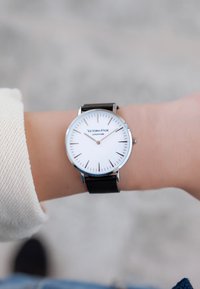 Orologio rotondo argento con quadrante bianco, bracciale a rete nera e marcatori dei minuti. Nome del marchio "Victoria-Hyde" in carattere blu al centro.