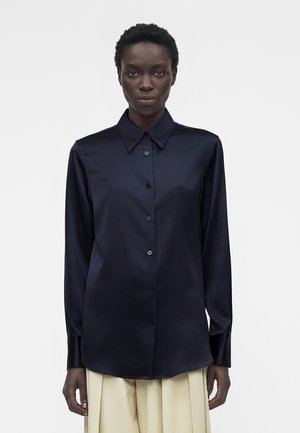 Femme portant une chemise noire en satin à manches longues boutonnée et un pantalon beige, debout devant un fond blanc uni.