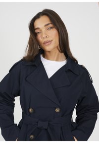 Granatowy trencz o podwójnym zapięciu, z dużym kołnierzem, paskiem w talii i guzikami; wykonany z gładkiego materiału z epoletem na ramionach.