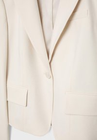 Cremfarbener Ein-Knopf-Blazer mit abgesteppten Revers und Pattentaschen, hängend vor weißem Hintergrund präsentiert.