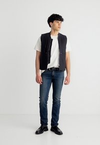 Νέος άνδρας που στέκεται, φοράει λευκό μπλουζάκι, μαύρο γιλέκο, μπλε τζιν, μαύρη ζώνη και μαύρα παπούτσια, κοιτάζοντας προς τα δεξιά του.