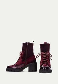 Un par de botines burdeos con tacones gruesos negros, cremalleras laterales y cordones delanteros, que combinan texturas de ante y cuero brillante.