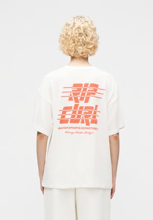 Άτομο με κοντά, σγουρά ξανθά μαλλιά, φορούσε ένα φαρδύ λευκό T-shirt με πορτοκαλί κείμενο "RIP CURL Water Sports Department" στην πλάτη.