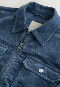 Veste en velours côtelé bleu avec col, fermeture éclair centrale et poches à rabat boutonnées, montrant la texture du tissu et l'étiquette de la marque à l'intérieur du col.