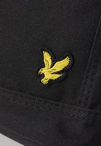 Svart teksturert stoff med brodert gult ørner logo. Sydd detaljer langs kantene forbedrer holdbarheten.