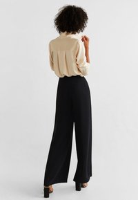 Beige bluse med detaljer i ryggen, sat sammen med sorte vide bukser. Glat tekstur, lange ærmer og en afslappet pasform.