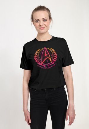Junge Frau mit hellem Haar, die ein schwarzes Starfleet Command T-Shirt und schwarze Hose trägt, steht vor einem schlichten hellen Hintergrund.