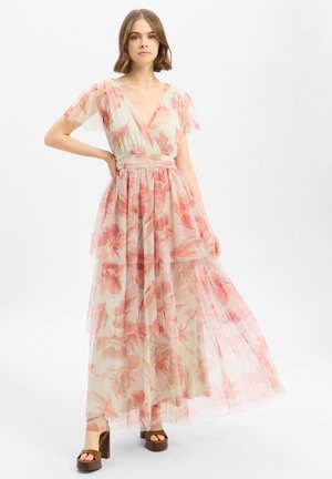 Jeune femme debout portant une longue robe fleurie transparente aux tons roses et pêche avec des talons compensés marron sur fond blanc.