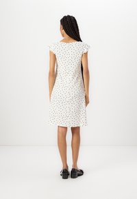 Valkoinen mekko mustilla pilkuilla, lyhyet hihat ja rento istuvuus. Ominaisuuksiin kuuluu polvimittainen helma ja teksturoitu kangas. Mustat kengät täydentävät asua.