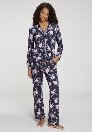 Femme aux cheveux bouclés portant un pyjama à manches longues bleu marine à fleurs avec des boutons, debout pieds nus avec une main dans la poche.