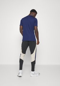 Marineblaues Baumwoll-T-Shirt kombiniert mit schwarz-beige Sporthosen, die seitliche Einsätze und Streifen aufweisen, getragen mit grauen Sportschuhen.