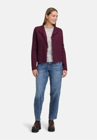 Burgunderfarbener Cardigan mit fransigen Kanten, kombiniert mit einem gestreiften Oberteil, dazu helle Jeans und braune Plateaustiefel.