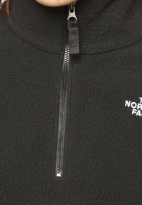 Primo piano della cerniera di una giacca in fleece nera con il logo parziale del marchio "The North Face" sul petto.