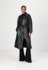 Čierny kožený trench coat s opaskom, širokými klopami a dlhými rukávmi. Má rovný strih a mierne otvorený predný diel.