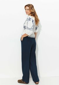 Blouse blanche brodée avec des motifs floraux bleus et des manches trois-quarts, associée à un pantalon large bleu marine et des chaussures à enfiler marron.