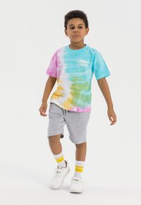 Majica s tie-dye vzorom v svetlo modrih, roza, rumenih in zelenih odtenkih, v kombinaciji s sivimi kratkimi hlačami, belimi športnimi copati in belimi nogavicami z rumenimi črtami.
