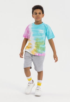 T-shirt tie-dye avec des motifs bleu clair, rose, jaune et vert, associé à un short gris, des baskets blanches et des chaussettes blanches avec des rayures jaunes.