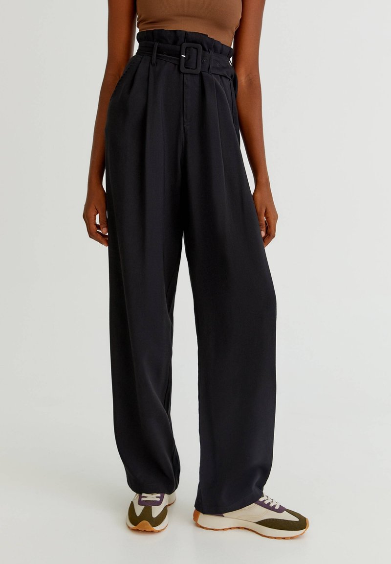 Personne portant un pantalon large taille haute noir avec une grande ceinture et des baskets dans des tons beige, vert olive et blanc.