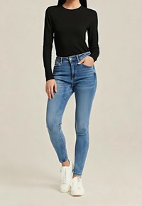 Femme portant un t-shirt noir à manches longues, un jean skinny taille haute bleu clair et des baskets blanches, debout avec une main dans la poche.