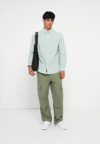 Hellgrün gestreiftes Hemd mit Knöpfen und einer Brusttasche, kombiniert mit olivgrünen Cargo-Hosen und weißen Sneakern. Schwarze Umhängetasche.